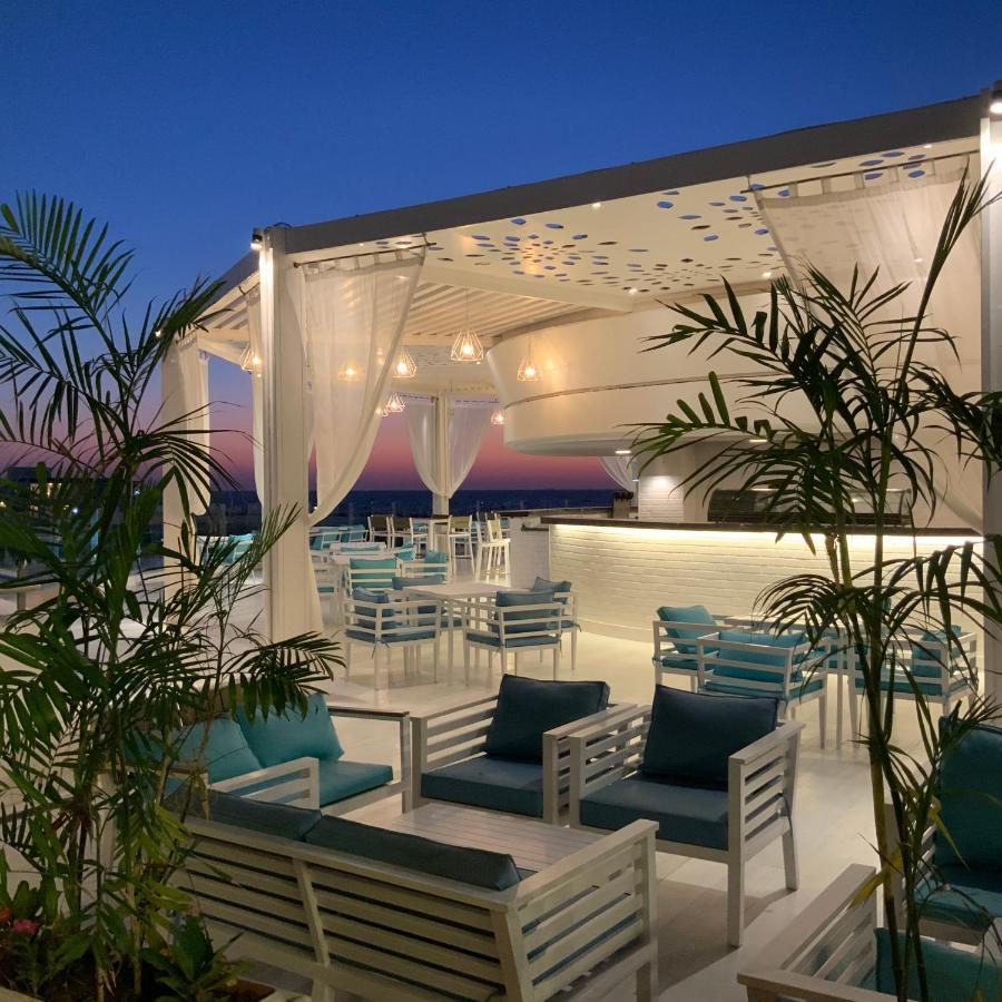 Sunrise Alex Avenue Hotel Αλεξάνδρεια Εξωτερικό φωτογραφία The photo shows a stylish outdoor terrace or rooftop lounge area. It features white furniture, including comfortable seating with teal cushions. The space is adorned with draped sheer curtains, providing a relaxed and elegant atmosphere. Lush green p