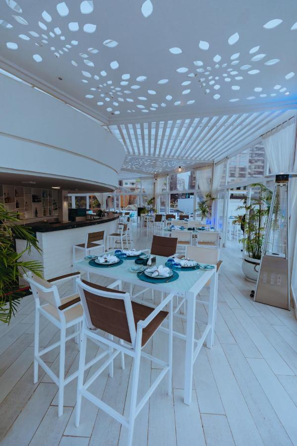 Sunrise Alex Avenue Hotel Αλεξάνδρεια Εξωτερικό φωτογραφία The photo shows a bright and modern restaurant interior. The space features white and light-colored furniture, including tables with a combination of white and brown chairs. Each table is elegantly set with plates, utensils, and decorative elements. 
