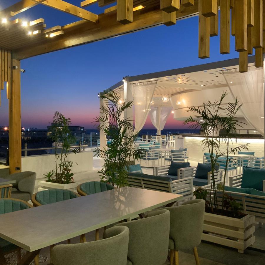 Sunrise Alex Avenue Hotel Αλεξάνδρεια Εξωτερικό φωτογραφία The photo shows a stylish outdoor dining area or terrace set against a beautiful sunset. There are several tables and chairs arranged neatly under a covered structure, with a backdrop of soft white curtains. The space is decorated with potted plants,