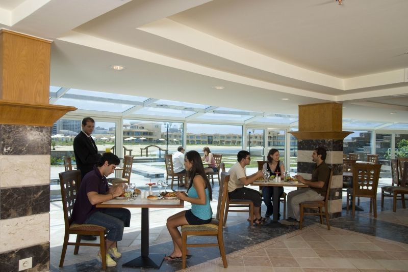 Sunrise Alex Avenue Hotel Αλεξάνδρεια Εστιατόριο φωτογραφία The photo shows an indoor dining area, possibly in a restaurant or café. There are several tables with people sitting at them. Two groups of diners are engaged in conversation while enjoying their meals. A server is also present, attending to one of 
