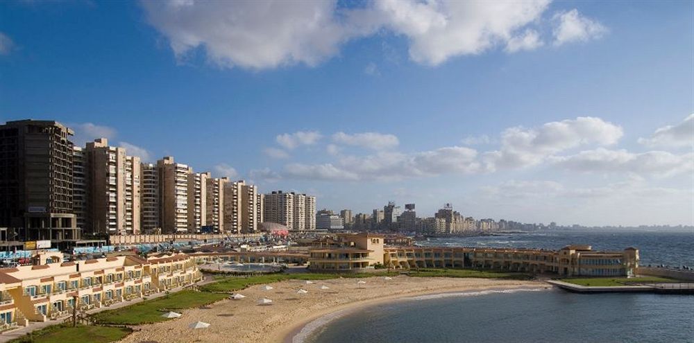 Sunrise Alex Avenue Hotel Αλεξάνδρεια Εξωτερικό φωτογραφία The photo shows a scenic coastal view featuring a sandy beach and a curved shoreline. In the foreground, there are several sun loungers and a landscaped area. Beyond the beach, there are multiple high-rise buildings lining the waterfront, indicating 