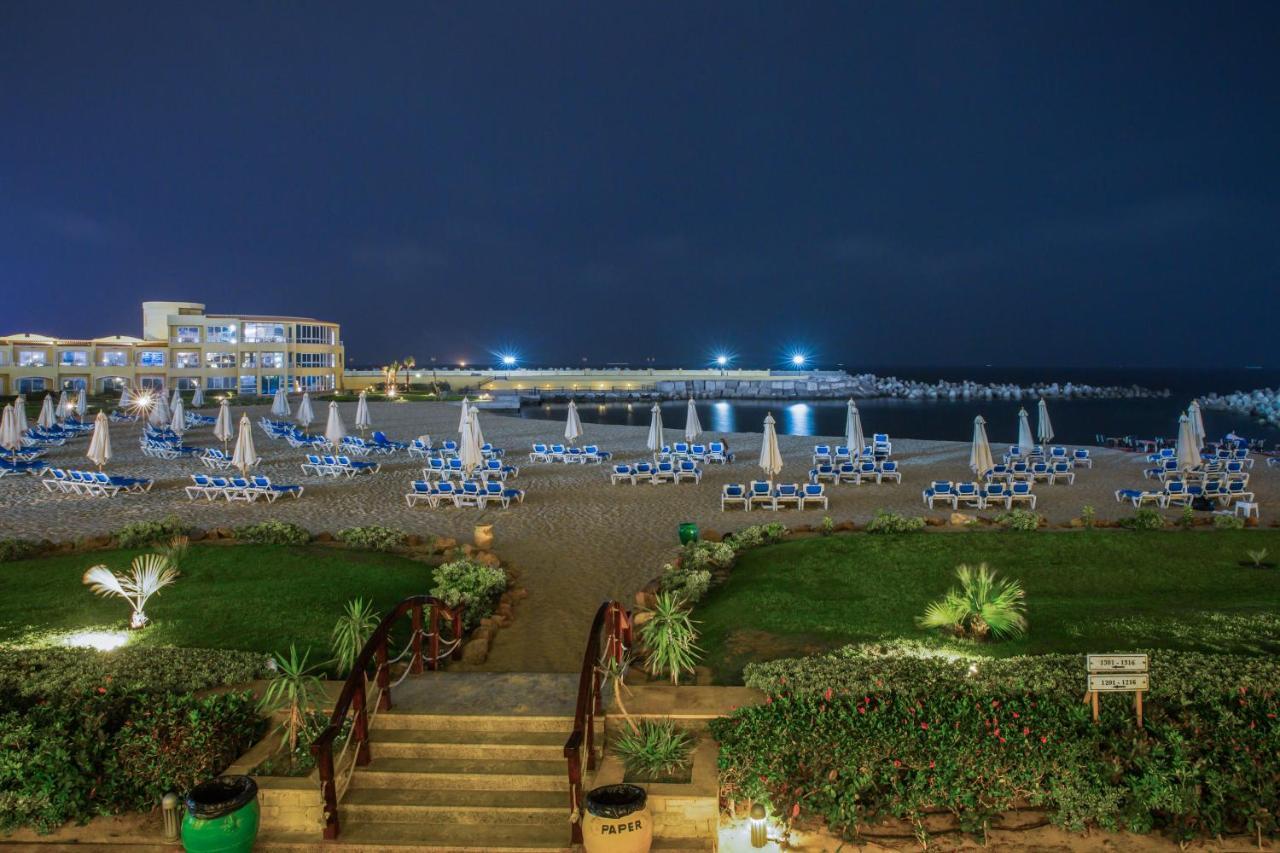 Sunrise Alex Avenue Hotel Αλεξάνδρεια Εξωτερικό φωτογραφία The photo shows a beach scene at night. In the foreground, there are steps leading down from a grassy area lined with plants. The beach is filled with numerous lounge chairs, all arranged with umbrellas, suggesting a relaxed atmosphere. In the backgr