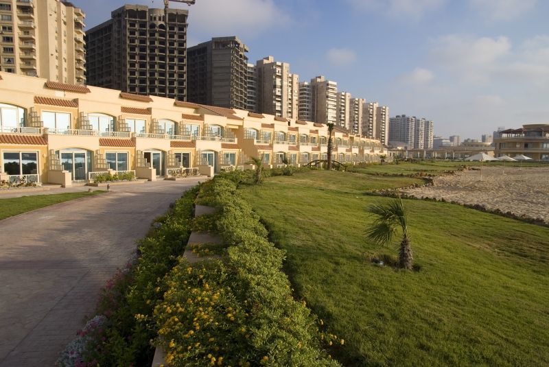 Sunrise Alex Avenue Hotel Αλεξάνδρεια Εξωτερικό φωτογραφία The photo depicts a scenic view of a shoreline area with modern buildings in the background. In the foreground, there is a well-maintained green lawn, bordered by a pathway. The buildings appear to be multi-story residential or commercial structures,
