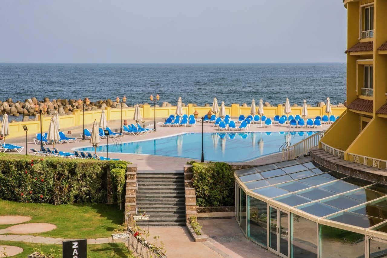 Sunrise Alex Avenue Hotel Αλεξάνδρεια Εξωτερικό φωτογραφία The photo depicts a scenic view of a pool area adjacent to the ocean. In the foreground, there is a large swimming pool surrounded by numerous blue lounge chairs and umbrellas, indicating a relaxing setting. There are steps leading down from a grassy