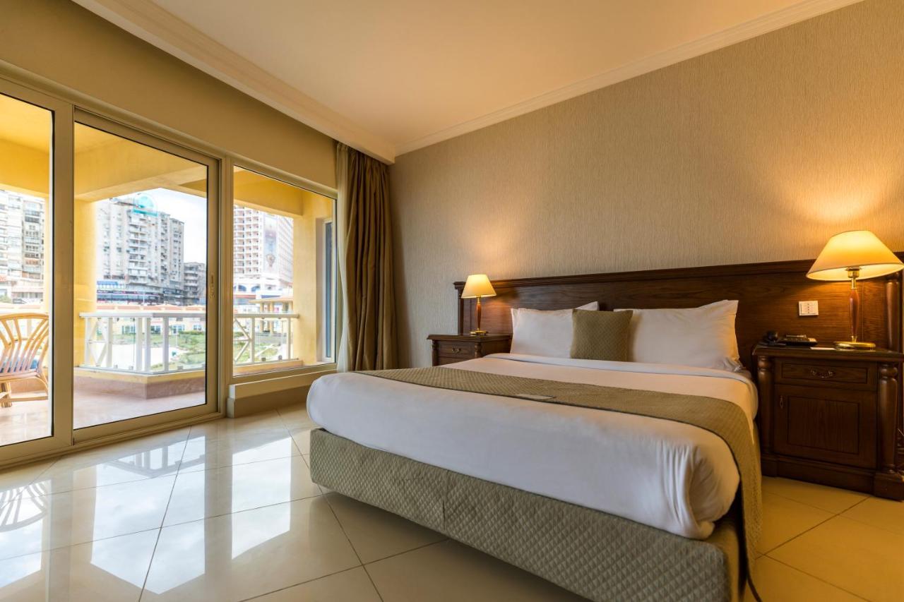 Sunrise Alex Avenue Hotel Αλεξάνδρεια Δωμάτιο φωτογραφία The photo shows a spacious hotel room featuring a large bed with white linens and decorative pillows. There are wooden nightstands with lamps on both sides of the bed. The walls are painted in neutral tones, and the floor is tiled. Large windows prov